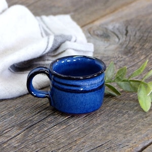 May include: A small, blue ceramic mug with a handle. The mug has a dark blue rim and three dark blue stripes around the middle.