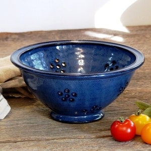 May include: A blue ceramic colander with a footed base and a pattern of small holes. The colander is sitting on a wooden surface with three red and yellow tomatoes.