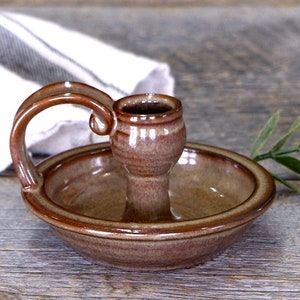 K&ouml;nnte beinhalten: Ein brauner Kerzenhalter aus Keramik mit einem einzelnen Kerzenhalter und einer kleinen Schale. Der Halter hat einen Griff und ist in Braun und Beige glasiert.