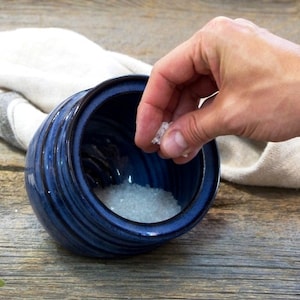 May include: A hand adds salt to a blue ceramic salt cellar. The salt cellar has a lid and is sitting on a wooden surface.