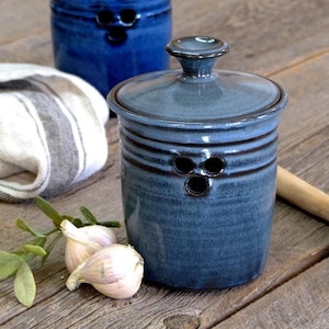 May include: A blue ceramic garlic keeper with a lid and three holes for ventilation. The keeper is sitting on a wooden surface with a wooden spoon and garlic cloves.