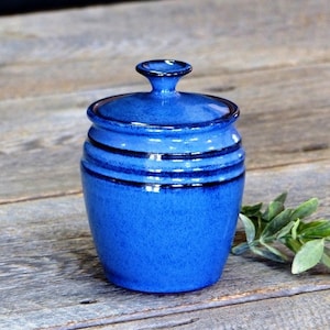 May include: A cobalt blue ceramic jar with a lid. The jar has a cylindrical shape with horizontal dark blue stripes. The lid has a small handle. The jar is on a wooden surface.