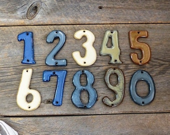 May include: A collection of ten ceramic house numbers, each with a unique color and glossy finish. The numbers range from 0 to 9 and are mounted on a rustic wooden surface. Each number has two pre-drilled holes for easy installation.