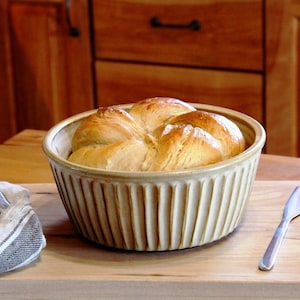 May include: A freshly baked loaf of bread in a beige ceramic baking dish with a ribbed design. The dish is sitting on a wooden chopping board with a butter knife and a pat of butter.