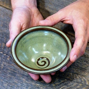 May include: A handmade ceramic bowl with a green glaze and a brown rim. The bowl has a spiral design in the centre. The bowl is held in two hands and is displayed on a wooden surface. This bowl is perfect for serving dips or small portions.