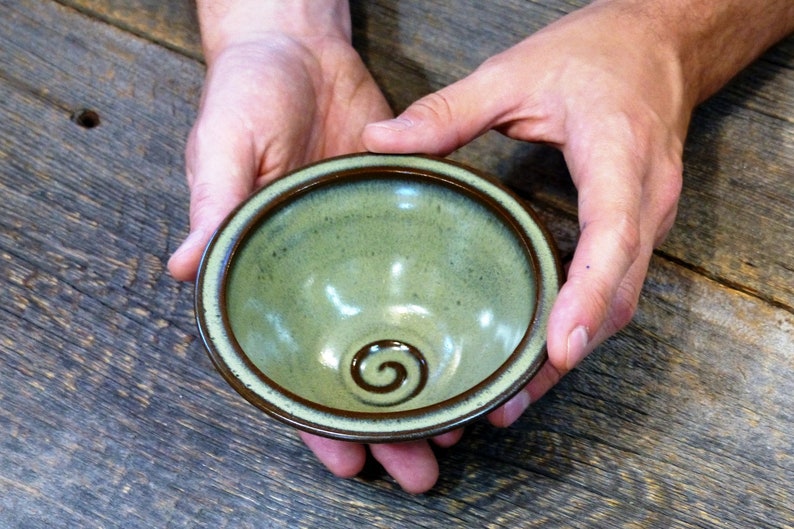 May include: A handmade ceramic bowl with a green glaze and a brown rim. The bowl has a spiral design in the centre. The bowl is held in two hands and is displayed on a wooden surface. This bowl is perfect for serving dips or small portions.