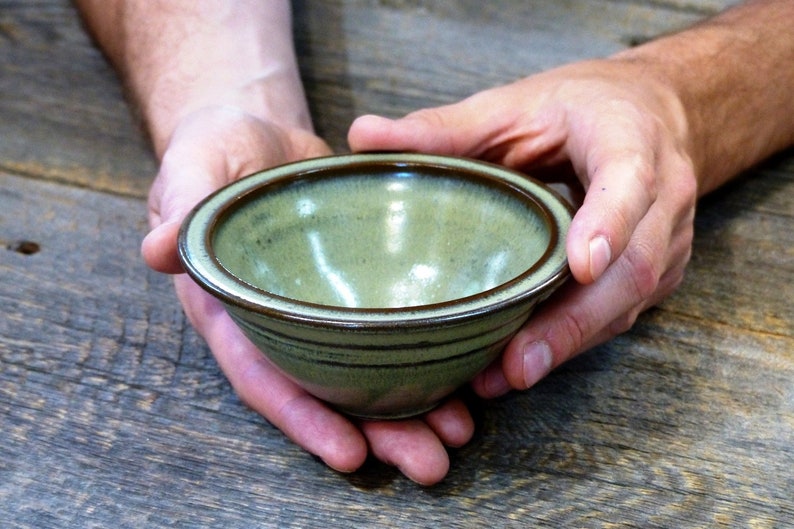 May include: A small, handmade ceramic bowl with a light green interior and a dark brown rim. The bowl has subtle horizontal lines and is held in two hands against a rustic wooden background. The bowl is approximately 5 cm tall and 13 cm in diameter.
