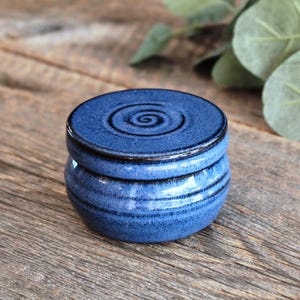 May include: A small, round, cobalt blue ceramic jar with a matching lid. The lid features a dark, hand-carved spiral design in the centre. The jar has a glossy, speckled glaze finish with dark blue accents around the rim and base, creating a textured appearance.