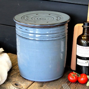 Puede incluir: Un recipiente de cer&aacute;mica azul con tapa y borde marr&oacute;n. El recipiente tiene una superficie texturizada y una abertura circular en la tapa. El recipiente est&aacute; sobre una superficie de madera.