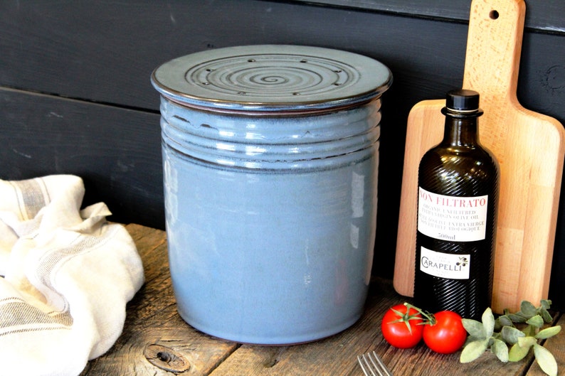 Puede incluir: Un recipiente de cer&aacute;mica azul con tapa y borde marr&oacute;n. El recipiente tiene una superficie texturizada y una abertura circular en la tapa. El recipiente est&aacute; sobre una superficie de madera.