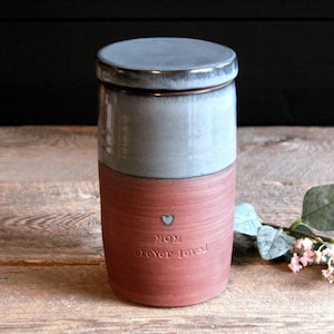 May include: A ceramic jar with a lid, painted in a light grey and a reddish brown colour. The jar has the words "Mom Forever Loved" and a small heart engraved on the side.