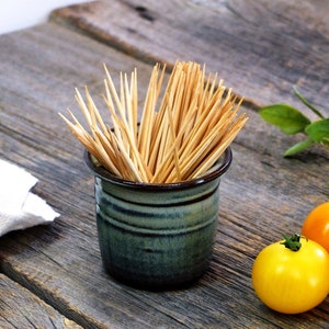 May include: A green ceramic toothpick holder filled with wooden toothpicks. The holder is sitting on a wooden table with three small tomatoes next to it.