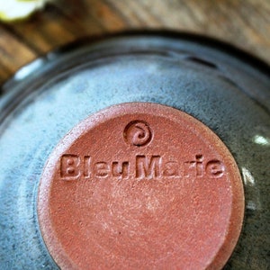 May include: A close-up shot of a ceramic bowl with a blue-grey glaze. The base of the bowl has a circular, reddish-brown stamp with the text "Bleu Marie" and a spiral design. The bowl is on a wooden surface.