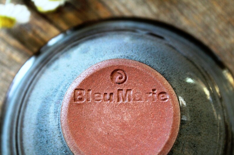 May include: A close-up shot of a ceramic bowl with a blue-grey glaze. The base of the bowl has a circular, reddish-brown stamp with the text "Bleu Marie" and a spiral design. The bowl is on a wooden surface.