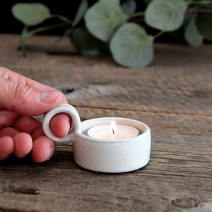 May include: A small, round, white ceramic candle holder with a single circular handle on the side. The holder features a smooth, glossy finish and contains a lit white tea light candle with a visible flame, creating a warm, steady glow.