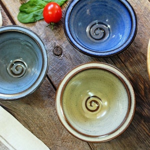 May include: Three ceramic bowls in blue, green, and grey, each with a spiral design in the centre. The bowls sit on a rustic wooden surface, with a wooden bowl and a tomato visible.