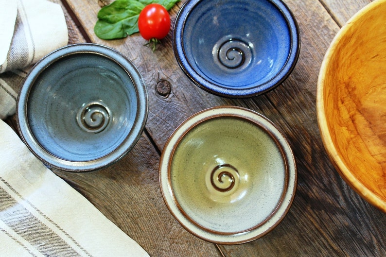 May include: Three ceramic bowls in blue, green, and grey, each with a spiral design in the centre. The bowls sit on a rustic wooden surface, with a wooden bowl and a tomato visible.