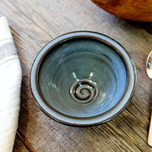 May include: A small, handmade ceramic bowl with a blue-green glaze and a spiral design in the centre. The bowl has a brown rim and sits on a wooden surface next to a silver spoon and a white cloth.