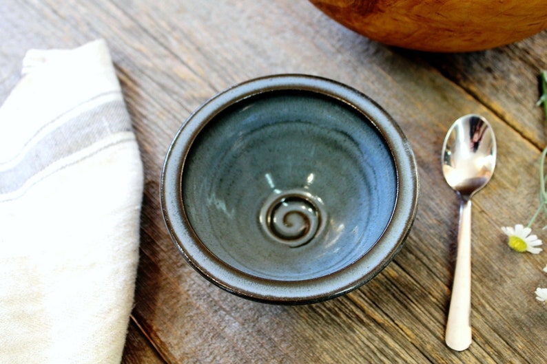 May include: A small, handmade ceramic bowl with a blue-green glaze and a spiral design in the centre. The bowl has a brown rim and sits on a wooden surface next to a silver spoon and a white cloth.