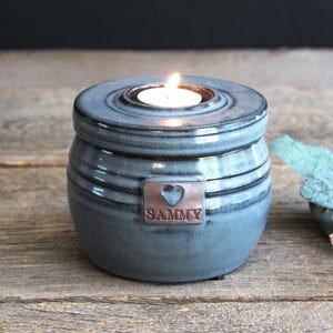 May include: A blue ceramic candle holder with a lit candle inside. The holder has a lid and a copper tag with a heart and the name "SAMMY". The candle holder is on a wooden surface.