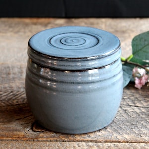 May include: A round, light blue ceramic jar with a matching lid. The lid features a circular, hand-carved spiral pattern on top. The jar has a smooth, glossy finish with dark blue accents along the rim and the lid edge, creating a subtle contrast.