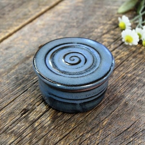 May include: A small, round, blue ceramic butter dish with a spiral design on the lid. The dish is sitting on a wooden surface.