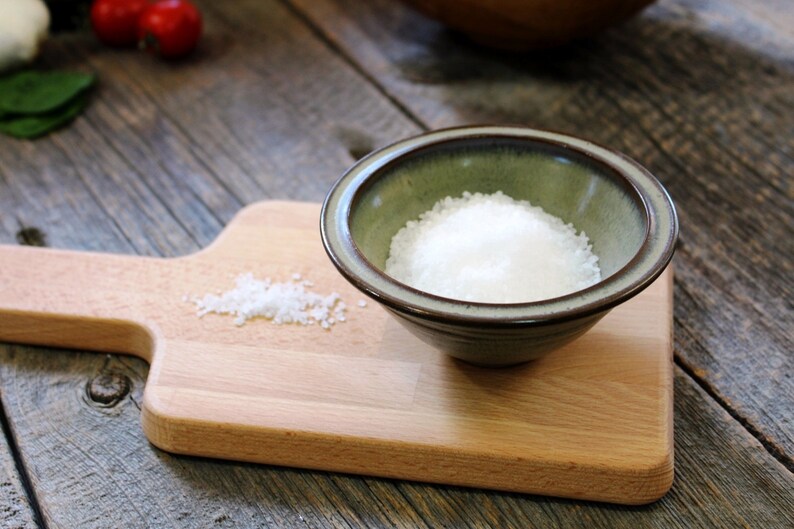 May include: A small, green ceramic bowl filled with white salt sits on a wooden chopping board. A small pile of salt is next to the bowl. The chopping board is on a weathered, grey wooden surface. The scene suggests a culinary theme.