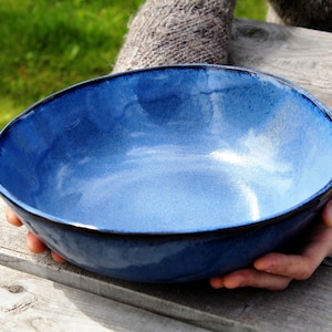 May include: A handmade, blue ceramic bowl with a glossy finish and a dark rim. The bowl is held by a person wearing a grey sweater. The bowl is sitting on a wooden surface, with green grass in the background. The bowl is approximately 25 cm in diameter.