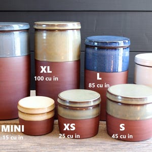 May include: Set of 8 ceramic storage jars in various sizes, ranging from mini to extra large. The jars have a two-toned design with a glazed top and a solid bottom. The jars are labeled with their size and volume in cubic inches: XXL 185 cu in, XL 100 cu in, L 85 cu in, M 65 cu in, S 45 cu in, XS 25 cu in, and Mini 15 cu in.