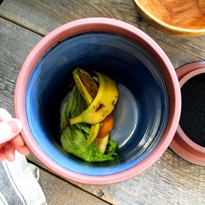 Puede incluir: Un contenedor de compost de cer&aacute;mica azul y rosa con una tapa negra. El contenedor contiene c&aacute;scaras de pl&aacute;tano y hojas de lechuga.