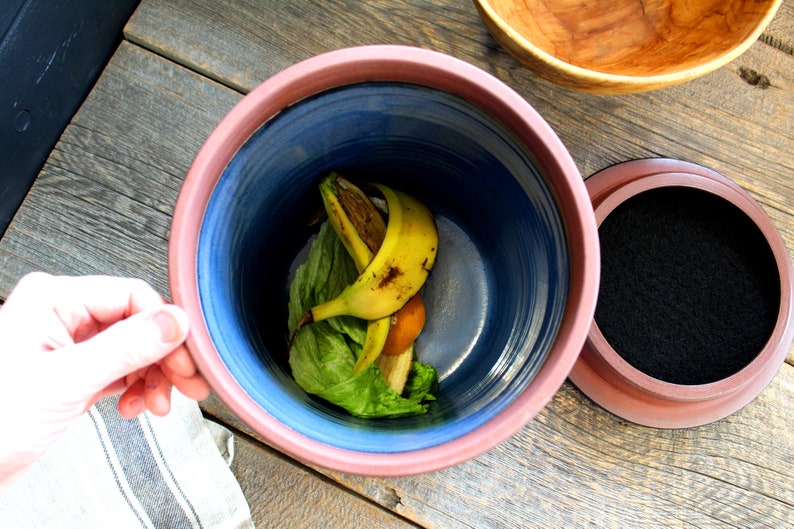 Puede incluir: Un contenedor de compost de cer&aacute;mica azul y rosa con una tapa negra. El contenedor contiene c&aacute;scaras de pl&aacute;tano y hojas de lechuga.
