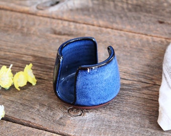 May include: A blue ceramic sponge holder with a dark blue glaze and a brown rim. The holder is shaped like a half-circle and has a small opening in the center.
