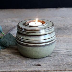 May include: A ceramic candle holder with a lit tea light. The holder is a muted green with brown horizontal stripes and a lid. The candle holder is on a weathered wooden surface.