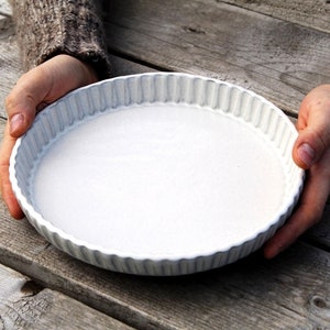 Peut inclure: Un plat à tarte en céramique blanche avec un bord cannelé. Le plat est tenu par deux mains.