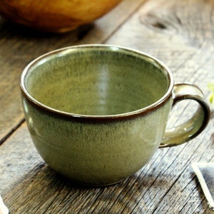Puede incluir: Una taza de t&eacute; de cer&aacute;mica verde con un borde marr&oacute;n y un asa. La taza est&aacute; sobre una superficie de madera.