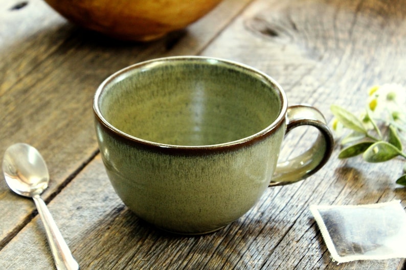 Puede incluir: Una taza de t&eacute; de cer&aacute;mica verde con un borde marr&oacute;n y un asa. La taza est&aacute; sobre una superficie de madera.