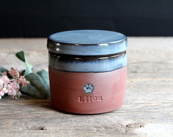 May include: A small, round, ceramic pet urn with a blue and brown glaze. The urn has a paw print and the name "Lilian" inscribed on the front.