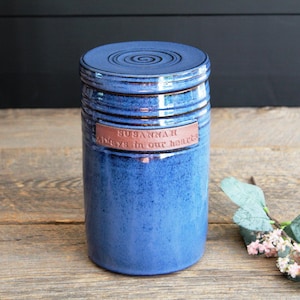 May include: A cylindrical blue ceramic urn with a matching lid featuring a spiral design. A rectangular terracotta-coloured plaque is attached to the front, embossed with the text "SUSANNAH" and "Always in our hearts" in capital letters.