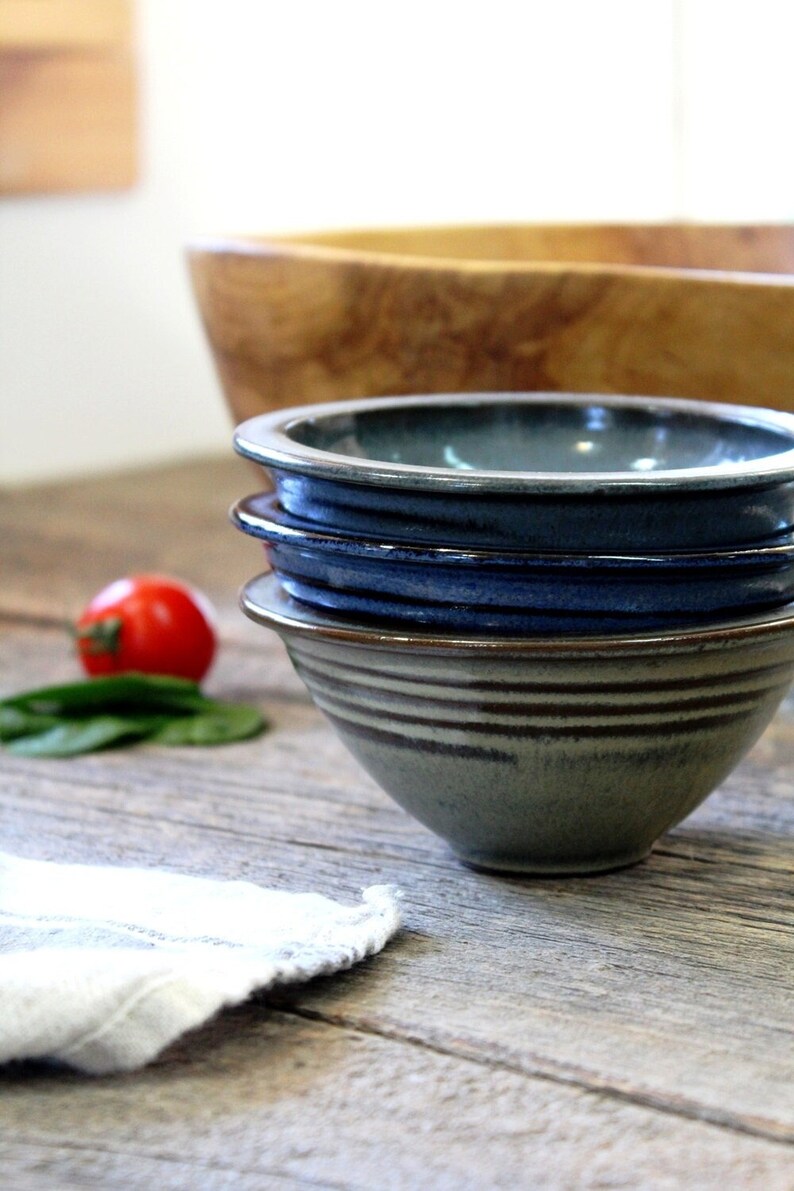 May include: Four ceramic bowls in shades of blue and green are stacked, with a wooden bowl in the background. The bowls have a textured, striped design. A red tomato and green leaves are on the wooden surface.