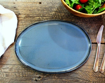 May include: A blue oval ceramic plate on a wooden table with a white cloth napkin, a wooden bowl with salad, and two silver utensils.