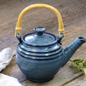 May include: A blue ceramic teapot with a wooden handle. The teapot has a rounded body and a lid with a knob on top. The teapot is sitting on a wooden surface.