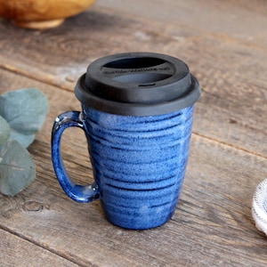 Peut inclure: Une tasse à café en céramique bleue avec un couvercle en silicone noir. La tasse a un design texturé et rayé.