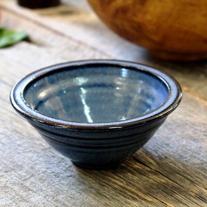 May include: A small, handmade ceramic bowl with a blue glaze and a brown rim. The bowl is round and has a slightly textured surface. It sits on a rustic wooden surface, suggesting a kitchen or dining setting.
