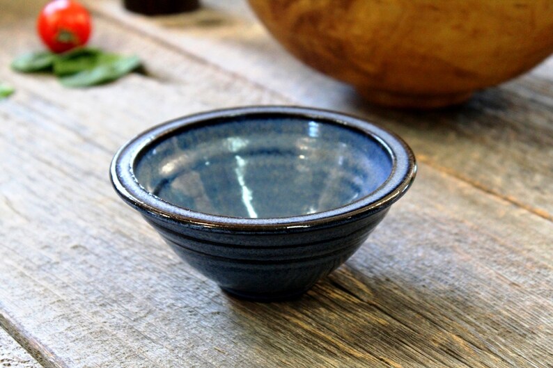 May include: A small, handmade ceramic bowl with a blue glaze and a brown rim. The bowl is round and has a slightly textured surface. It sits on a rustic wooden surface, suggesting a kitchen or dining setting.
