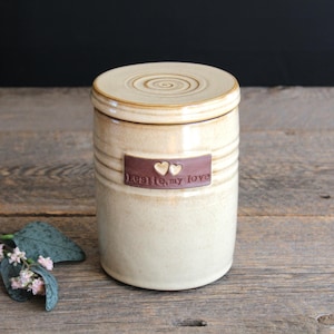 May include: A cylindrical ceramic jar with a glossy cream-coloured glaze and a matching lid featuring a spiral pattern. A rectangular brown clay plaque is attached to the front, embossed with two small hearts and the text "Leslie, my love."