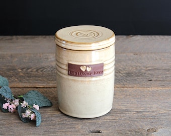 May include: A cylindrical ceramic jar with a glossy cream-colored glaze and a matching lid featuring a spiral pattern. A rectangular brown clay plaque is attached to the front, embossed with two small hearts and the text "Leslie, my love."