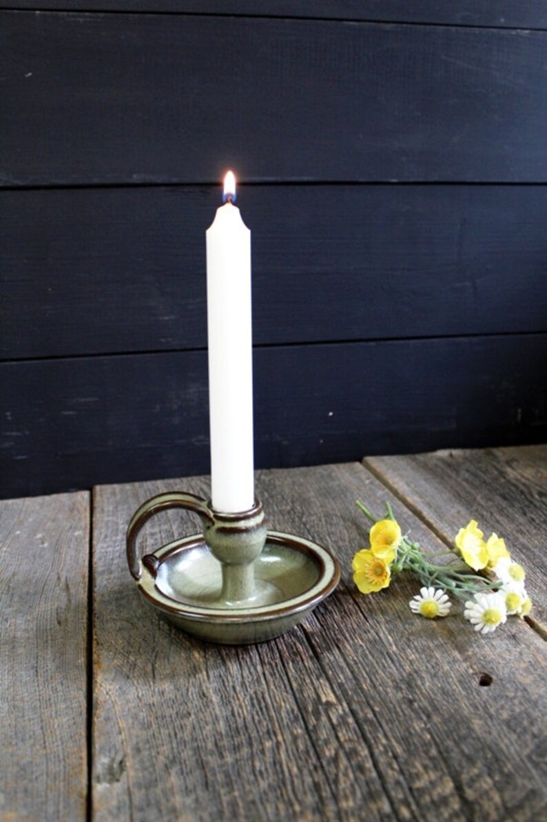 K&ouml;nnte beinhalten: Ein gr&uuml;ner Kerzenhalter aus Keramik mit einem Griff und einer brennenden wei&szlig;en Kerze. Der Kerzenhalter steht auf einer Holzoberfl&auml;che mit gelben und wei&szlig;en Blumen.