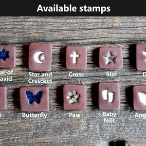 May include: A collection of 10 ceramic tiles with different designs, including a star of David, a star and crescent, a cross, a star, a dove, a heart, a butterfly, a paw print, baby feet, and angel wings. The tiles are all brown with white or gold designs.