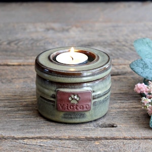 May include: A ceramic candle holder with a lit tea light. The holder is green and brown with a rectangular plaque that reads "Victor" and features a paw print. The candle holder is on a wooden surface.