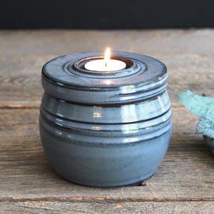 May include: A blue ceramic candle holder with a lit tea light. The holder has a round shape with horizontal ridges and a lid. The candle holder is on a wooden surface.